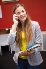 Female worker wearing gray blazer holding smartphone, folder, standing in red accent wall office