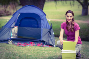 Female camper crouching on grass at campsite opening cooler beside dome tent and mug, copy space