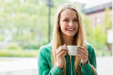 Smiling teenage girl wearing green zip-up sweatshirt standing on cafe patio holding white mug