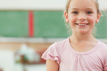 Female child student standing in classroom in front of green chalkboard with red stripe, copy space