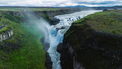 Gullfoss waterfalls tumble dramatically into a verdant canyon in Iceland, surrounded by lush...