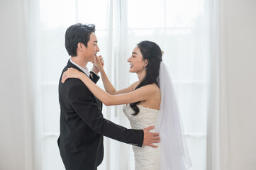 Happy Asian couple in wedding attire, hugging each other and smiling joyfully. The bride is wearing...