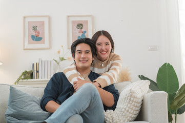 Happy young Asian couple hugging, sitting on cozy couch together, overjoyed laughing woman and man...