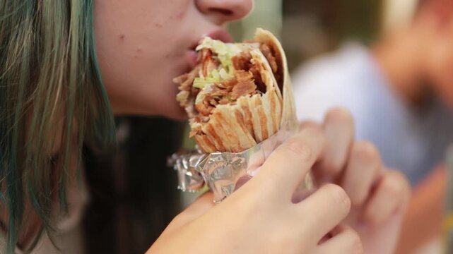 Young woman with partially teal-dyed hair savors a doner kebab wrapped in foil, taking bites and enjoying the flavors in an outdoor setting