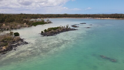 Drone shot of the tranquil north coast of Mauritius showing turquoise waters, volcanic rock islets, and lush coastal vegetation under a bright sky.