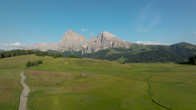 Aerial view of green fields with wooden structures and mountains under a clear blue sky, creating a vibrant contrast of colors and textures, Compatsch, Trentino-Alto Adige, Italy.