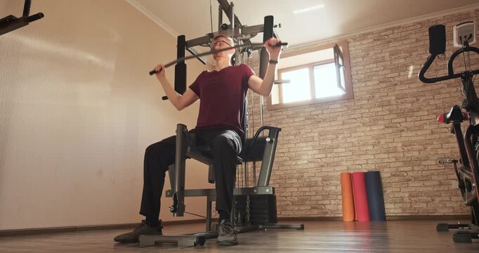 Athletic man doing lat pulldown exercise on home gym machine in modern apartment, open window and yoga mats. Healthy active lifestyle, strength training, workout routine, fitness motivation