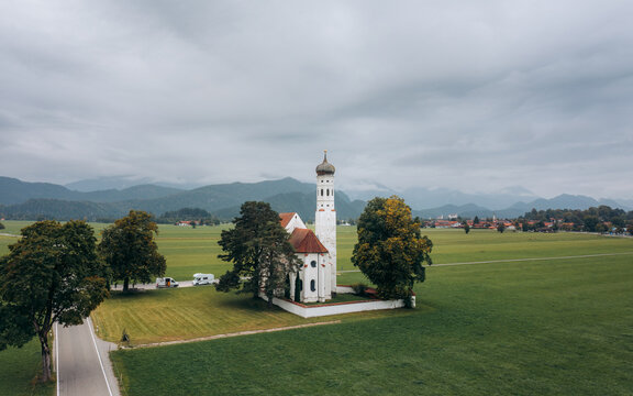 Aerial view of the striking white St. Coloman Church amidst verdant fields, framed by distant mountains under a serene sky, Schwangau, Bavaria, Germany.