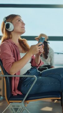 A woman listens to music while waiting at the airport. Other travelers are visible in the background.