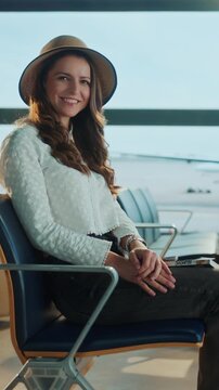 A woman in a hat smiles while waiting at an airport terminal. Others are visible in the background, including a family and elderly person.