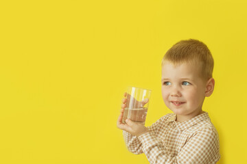 A smiling child holds a glass of fresh drinking water on a yellow background. Drinking water for children.