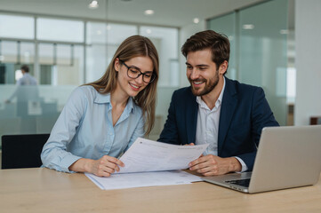 Obraz premium Two smiling business professionals reviewing documents together at a desk with a laptop
