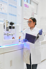 Focused young latina researcher with her hair tied back, dressed in a white lab coat and glasses, works on a 3D printer in a modern academic laboratory environment