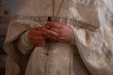 Orthodox priest in ornate white vestments holds a small bottle and a liturgical brush during a...