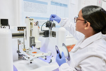 Scientist woman feeds a new spool of filament into a lab-grade 3D printer during setup, wearing a lab coat and working with care