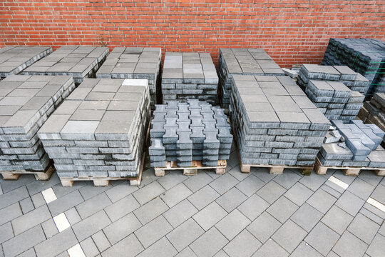 Konzept Baustelle Vorbereitung, Lieferung, Pause oder Baustopp: Unterschiedliche Pflastersteine auf Paletten gestapelt auf einem Grundst&uuml;ck vor einer roten Klinkerwand &ndash; selektiver Fokus, Textfreiraum
