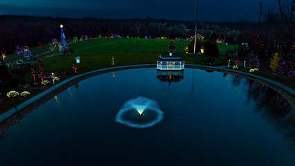 Colorful holiday lights reflect off the pond's surface as a fountain adds a shimmering display. Festive decorations surround the park, creating a magical winter atmosphere after dusk.