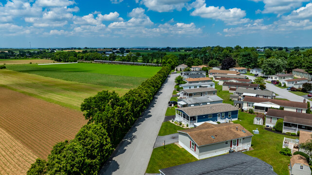 Aerial view of a peaceful residential Mobile, Manufactured Homes community surrounded by lush green fields and vibrant blue sky. Homes line a well-maintained road, showcasing quiet suburban lifestyle