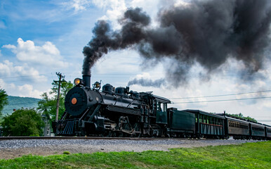 A historic narrow gauge steam locomotive moves along the tracks in a rural area, releasing black smoke into the clear blue sky. Lush greenery surrounds the railway, creating a picturesque setting.