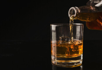 Pouring whiskey from bottle into glass with ice cubes on black background, copy space