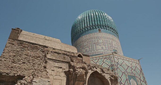 Ancient ruins of brick entrance wall in front of turquoise dome and tall minaret of Gur e Amir Mausoleum in Samarkand, Uzbekistan under blue sky, showcasing Islamic architecture, restoration contrast