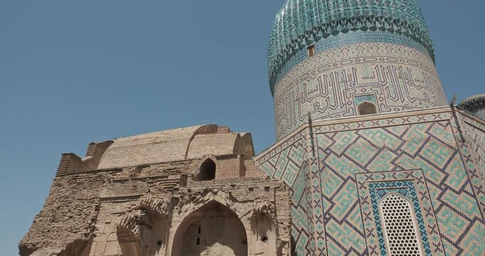 Ancient ruins of brick entrance wall in front of turquoise dome and tall minaret of Gur e Amir Mausoleum in Samarkand, Uzbekistan under blue sky, showcasing Islamic architecture, restoration contrast