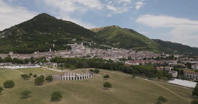 Aerial view of Gubbio, showcasing the ancient Roman theater ruins amidst the green landscape, with the town nestled against the backdrop of mountains, Gubbio, Perugia, Italy.