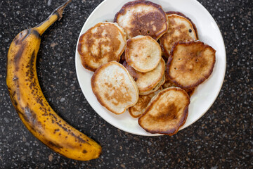 Ripe banana next to plate of pancakes on black tabletop. Banana pancakes for breakfast, top view