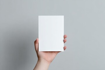 A hand holding a single blank white A5 vertical flyer or invitation card in front of a soft gray background, centered composition, realistic studio lighting