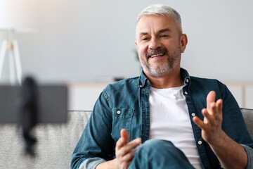 Happy handsome senior man vlogger or blogger broadcasting from home, sitting on couch in cozy...