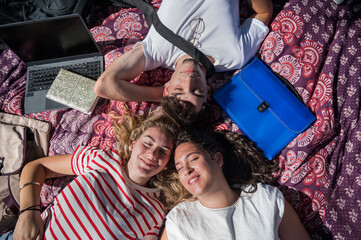 Three university students lying on a blanket outdoors with eyes closed, smiling and relaxing...