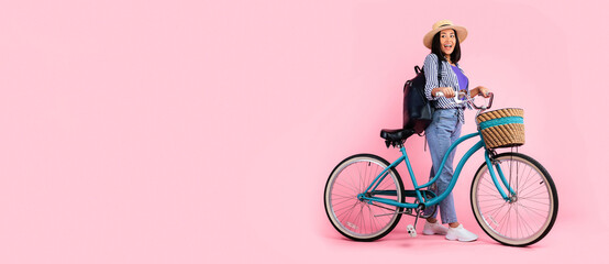 Obraz premium Full length portrait of cute excited asian woman wearing summer hat and backpack standing with classic bicycle with wicker basket looking back at free copy space isolated on pink studio background