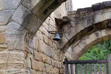 Bell suspended beneath a stone arch, framed by weathered masonry and soft greenery—quiet echoes...
