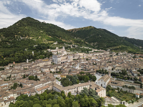 Aerial view of terracotta rooftops cascading down the slopes towards the Palazzo dei Consoli, nestled amidst lush greenery under a vast sky, Gubbio, Perugia Province, Italy.