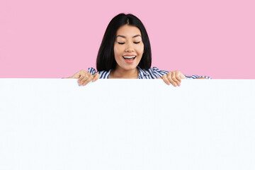 Place For Your Promo. Excited asian woman standing behind white empty board for advertisement over pink background. Lady peeking out big blank advertisement panel, looking down at empty space for text © Prostock-studio
