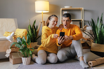 A surprised couple sits on the floor of their new home, looking at a smartphone with wide eyes and open mouths, surrounded by moving boxes and a houseplant.