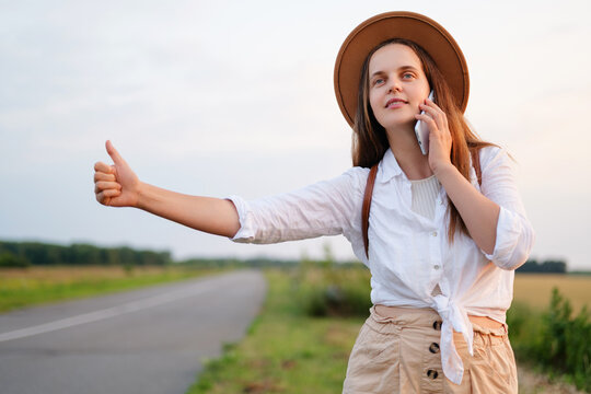 Young woman in a hat hitchhiking with her thumb out, while also talking on her smartphone, on a rural road. - Powered by Adobe