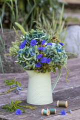 Blumenstrauß mit blauen Hortensienblüten, Federnelken, Wilder Möhre, Gräsern und Brombeerzweigen im vintage Krug im Garten