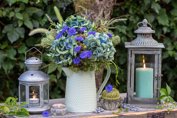 Garten-Arrangement mit Blumenstrauß mit blauen Hortensienblüten, Federnelken, Wilder Möhre, Gräsern und Brombeerzweigen und vintage Laternen