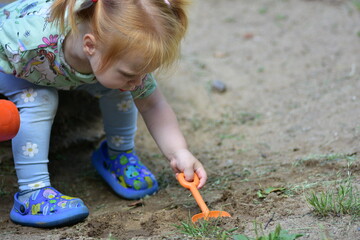 Red-haired toddler girl crouches and digs into the ground with a small orange shovel in a sandbox area, fully focused on her playful outdoor activity in natural surroundings. © Karolina