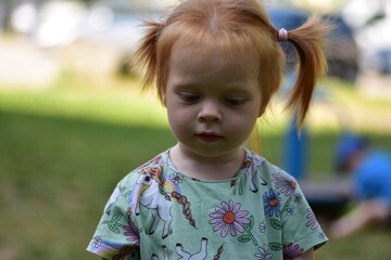 Red-haired toddler girl with pigtails wearing a unicorn shirt looks down thoughtfully in an outdoor...