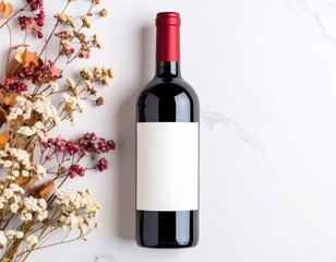 A bottle of red wine with a blank label and dried flowers on a white marble surface top down view