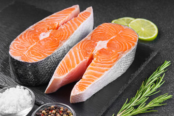 Two fresh raw salmon or trout steaks on black stone surface with food ingredients. Lime, rosemary,...