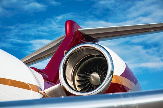 Close-up of a private jet engine with detailed turbine blades, mounted on a red and white fuselage. The image shows part of the T-tail, shot in natural front light under a bright blue sky. 