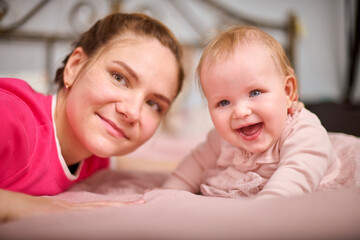 Young mother with brown hair, smiling baby girl indoors. Warm pink tones, soft lighting, metal...