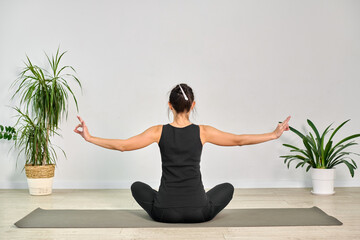 Young woman practices yoga in serene, minimalist room. She sits cross-legged on mat with arms...