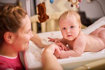 Smiling adult mother with brown hair plays with joyful baby girl on changing table. Soft lighting...