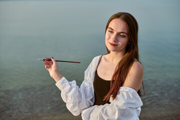 Young adult female painter stands by calm lake, holding paintbrush. Her expression reflects...