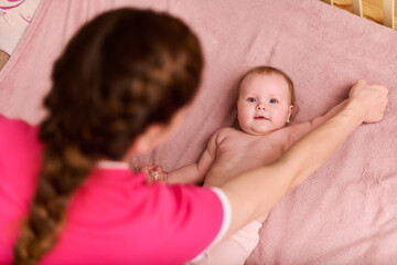 Young mother helping baby girl with passive exercises. Baby lying on pink blanket, relaxed...