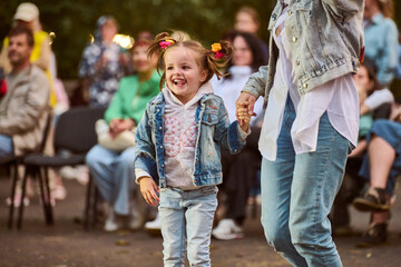 Young girl with pigtails and colorful clips, wearing denim, dances happily at outdoor gathering....